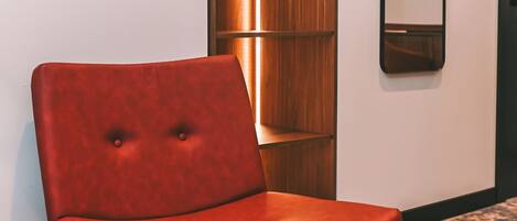 A red upholstered chair with black legs in a hallway with a patterned carpet, a wooden shelving unit, and a mirror on the wall.