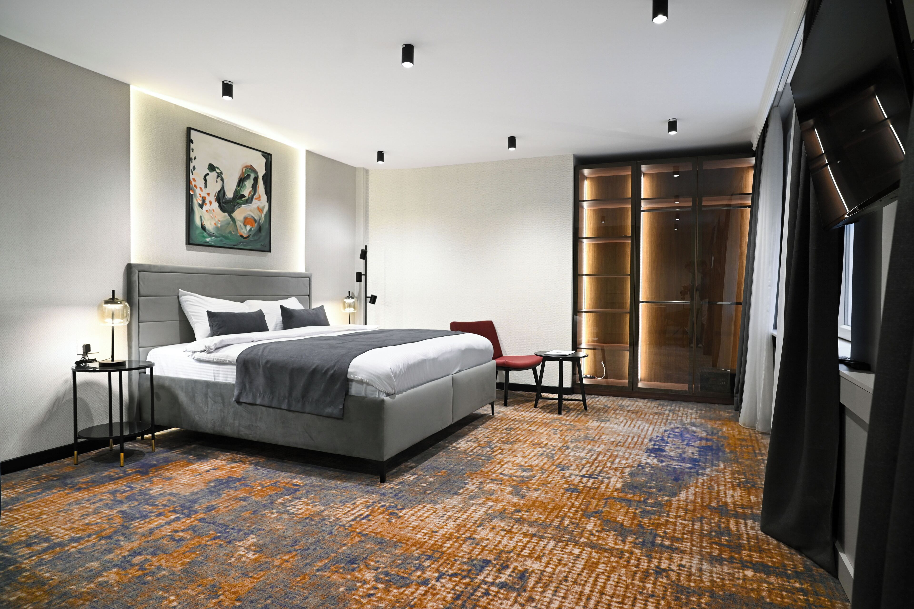 Une chambre d’hôtel moderne avec un grand lit, un fauteuil rouge, une petite table et une porte vitrée donnant sur un balcon.