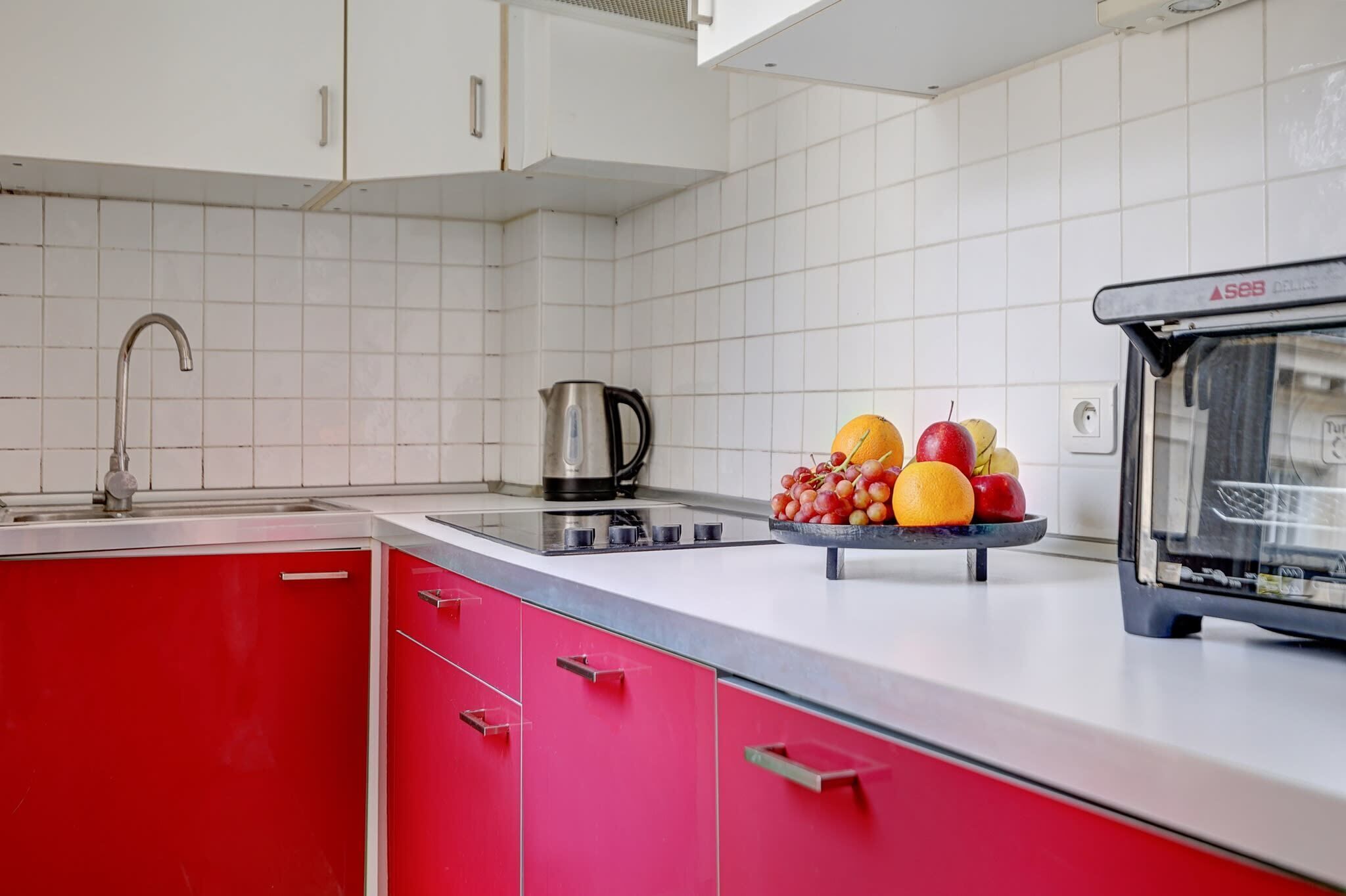 Une cuisine moderne avec des murs recouverts de carreaux blancs, un meuble rouge, un évier, une bouilloire et un panier de fruits posé sur le plan de travail.