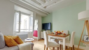 A modern living room with a sofa, dining table, and chairs. A television is mounted on the wall, and a window provides natural light.