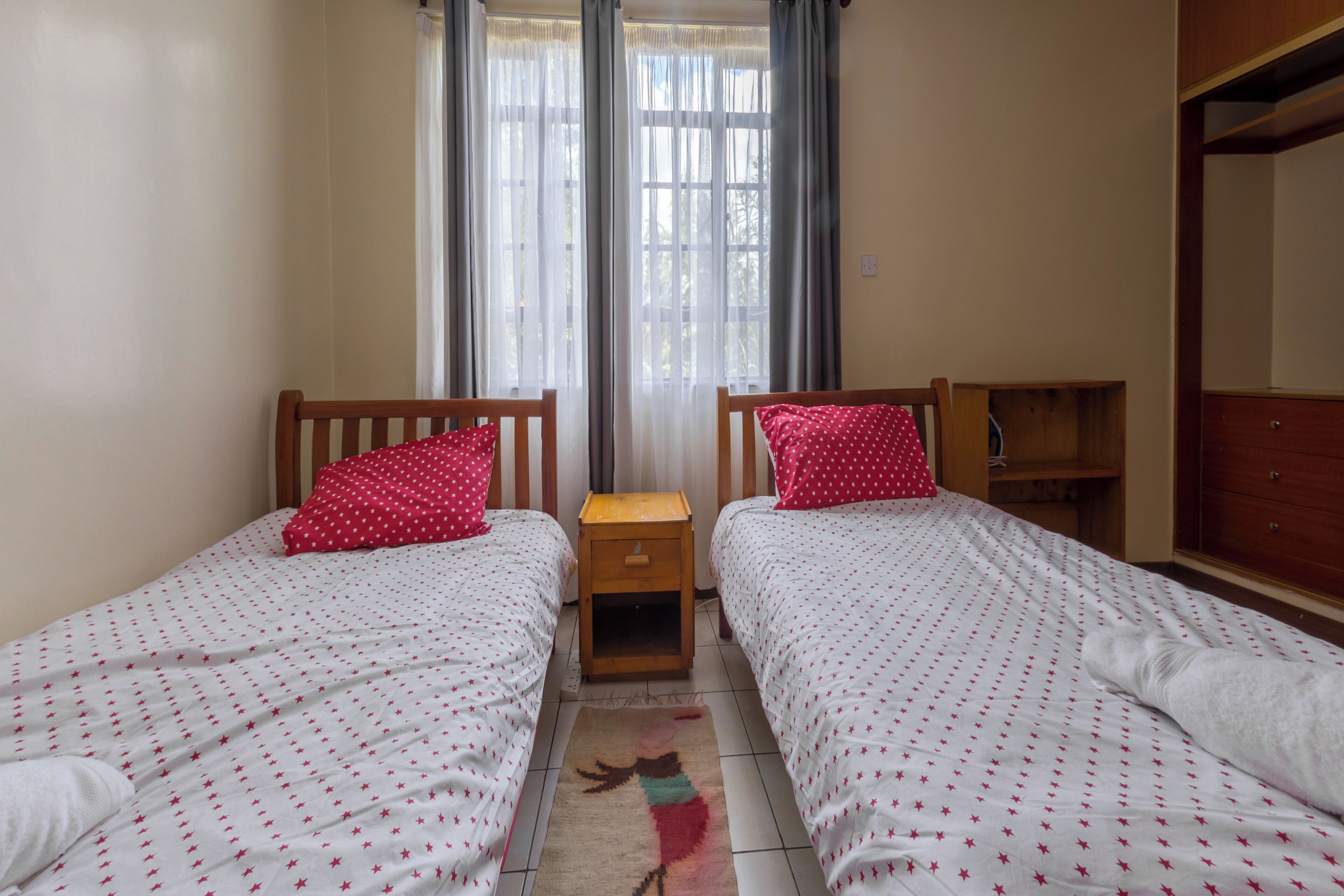 Une chambre à deux lits, chacun étant agrémenté d’un oreiller rouge, d’une petite table de chevet en bois et d’une fenêtre avec des rideaux.