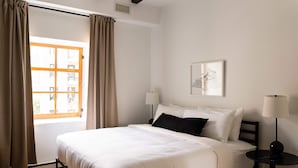 A neatly made bed with white linens, a bedside table with a lamp, and a window with curtains. - Maison Rasco • One-Bedroom Apartment • King Size Bed • Old Montreal • Full Kitchen • Stone Wall • Pet Friendly • Self Check-In Access • Wi-Fi • AC (Montreal)