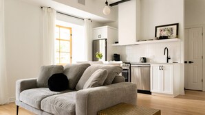 A modern living room with a grey sofa, a small wooden table, and a kitchen area with white cabinets and appliances. - Maison Rasco • One-Bedroom Apartment • King Size Bed • Old Montreal • Full Kitchen • Stone Wall • Pet Friendly • Self Check-In Access • Wi-Fi • AC (Montreal)