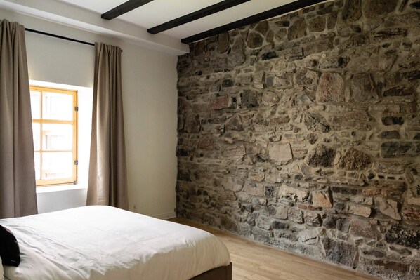 A room with a stone wall, a wooden floor, a bed with a white bedspread, and a window with curtains.