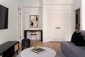 A modern living room with a grey sofa, a round coffee table with a book and flowers, a black and white patterned ottoman, a black and white framed abstract painting, a white door, and a white wall-mounted TV.