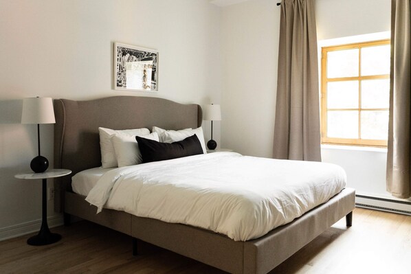 A neatly made bed with white linens, two bedside lamps, a framed picture on the wall, and a window with curtains.