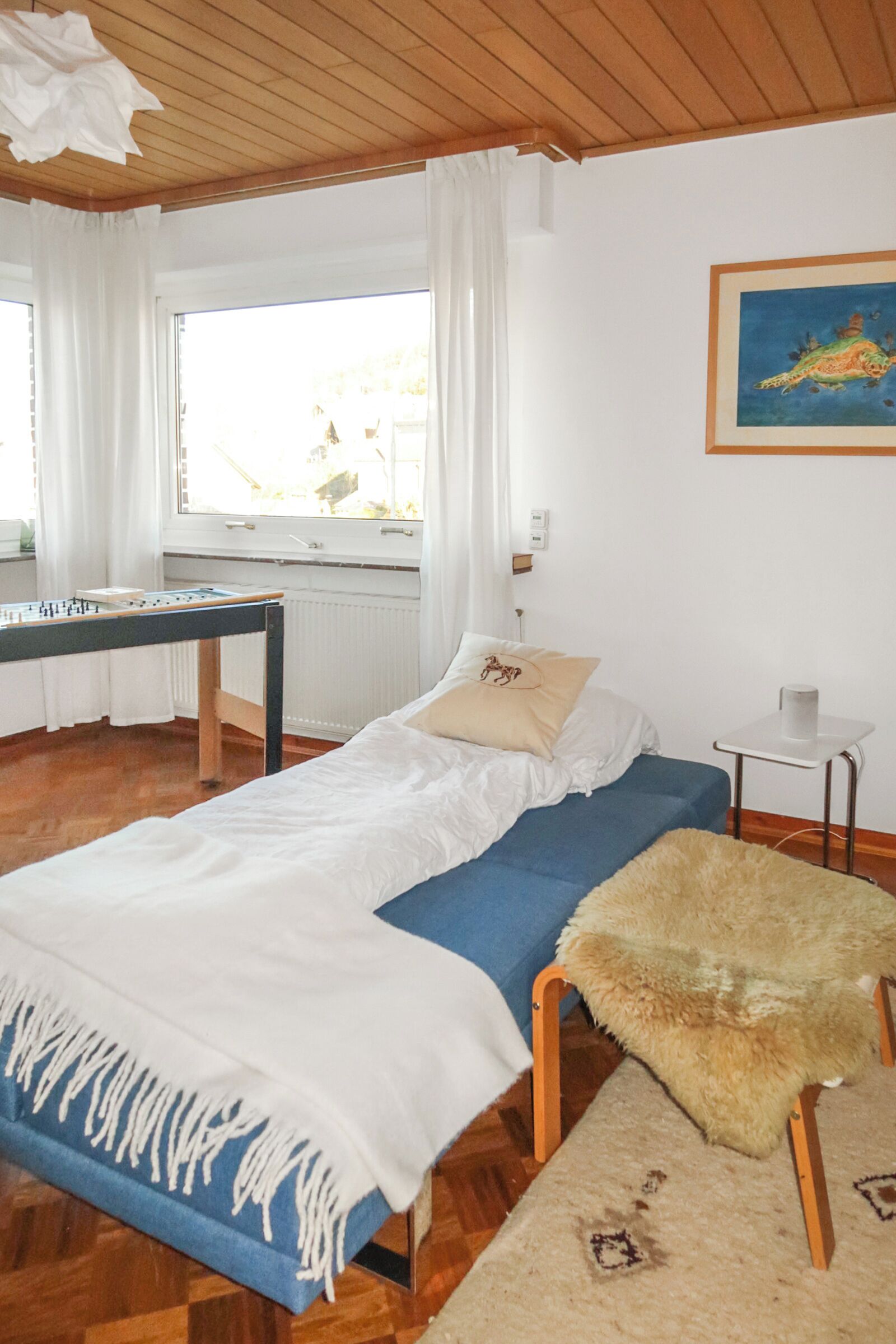 A room with a bed, a small table, a window with curtains, and a painting on the wall.