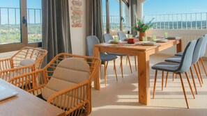 Un coin repas avec des chaises en osier, une table en bois et une vue sur le paysage grĂące Ă de grandes fenĂȘtres.