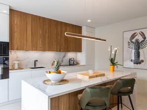 A modern kitchen with wooden cabinets, a marble countertop, a central island with bar stools, and a framed lobster print on the wall. - Charm & Serenity with Private Terrace and Garden View (Saint-Martin)