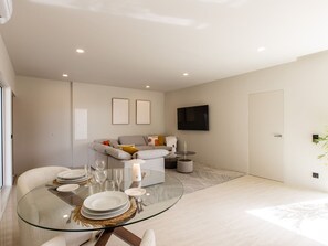 A modern living room with a glass dining table set for four, a grey sofa, and a flat-screen TV mounted on the wall. - Charm & Serenity with Private Terrace and Garden View (Saint-Martin)
