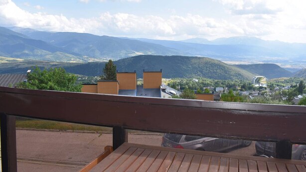 Blick vom Balkon mit Holzgeländer auf ein Tal, in dem sich Berge in der Ferne zeigen, sowie einige Gebäude und ein klarer Himmel.