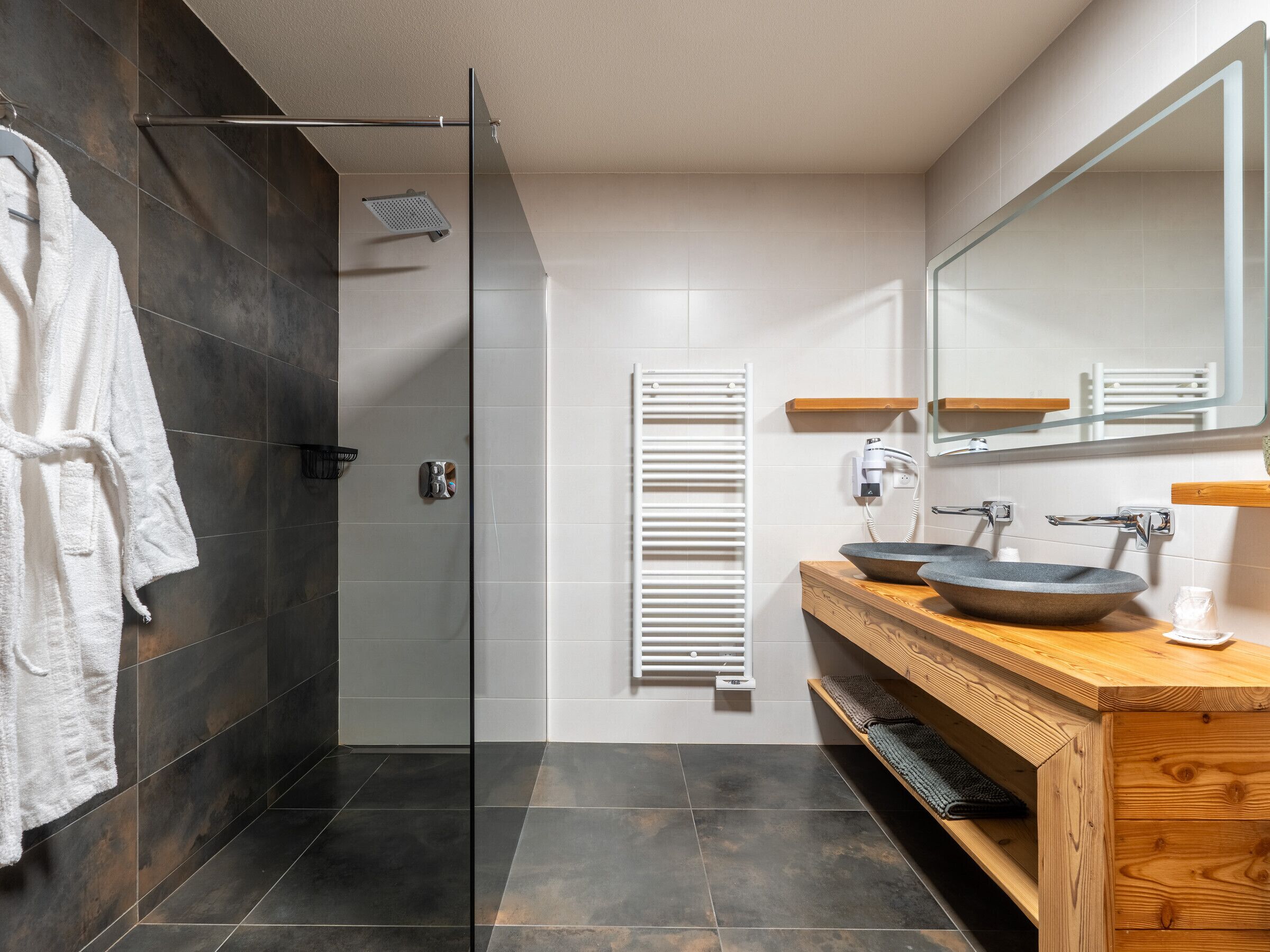 Uma casa de banho moderna com cabine de chuveiro em vidro, porta-toalhas branco, toucador de madeira com lavatório suspenso e um espelho grande.
