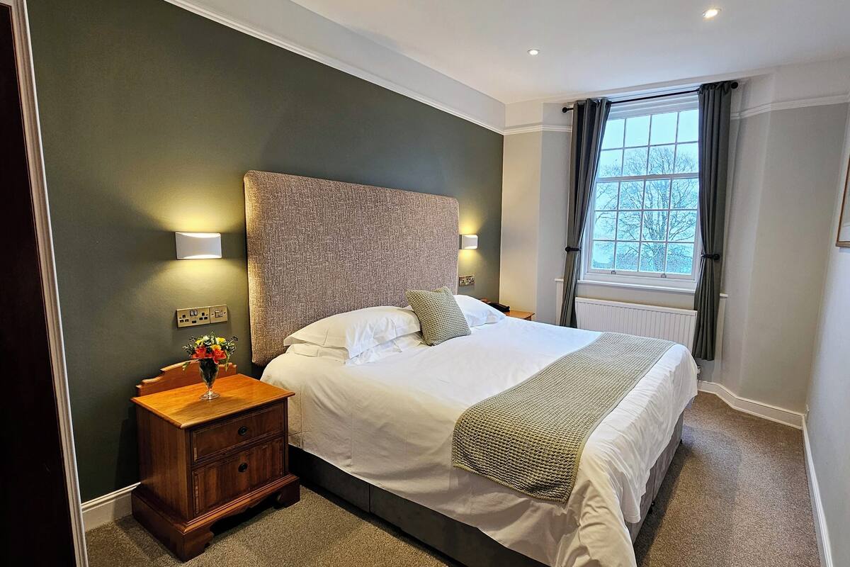 Ein ordentlich eingerichtetes Hotelzimmer mit einem großen Bett, einem Nachttisch mit Blumenvase, einem Fenster mit Vorhängen und einer Wandleuchte.