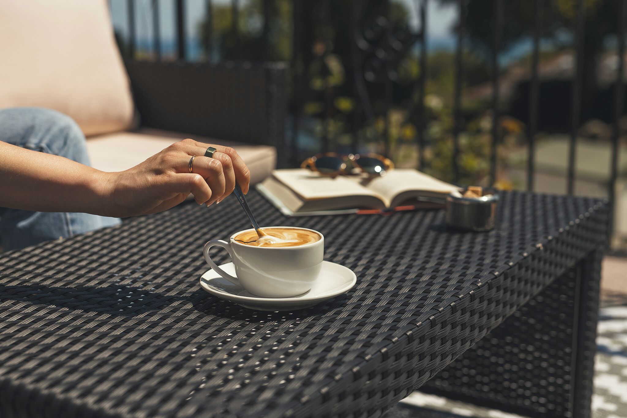 Une personne assise à une table, avec une tasse de café, un livre et des lunettes de soleil.