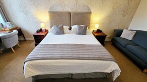 Une chambre d’hôtel avec un grand lit, deux lampes de chevet, un bureau avec une chaise, un canapé et un mur orné d’un motif représentant un arbre.