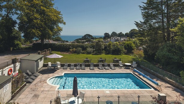 A rectangular swimming pool with lounge chairs and umbrellas, surrounded by a fence and greenery, overlooking a coastal landscape. - Stoke Lodge Hotel (Dartmouth)