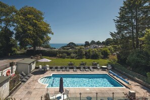 Piscina rectangular con sillas de descanso y sombrillas, rodeada por una valla y follaje, con vistas a un paisaje costero.