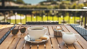 A table with a cup of coffee, sunglasses, and a sugar bowl on a wooden deck with a view of the ocean. - Stoke Lodge Hotel (Dartmouth)