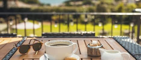 A table with a cup of coffee, sunglasses, and a sugar bowl on a wooden deck with a view of the ocean.