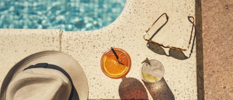 A hat, sunglasses, and two drinks on a sun lounger by a pool.