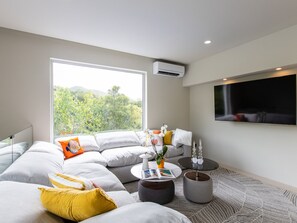 A modern living room with a large window, a sectional sofa, a coffee table, and a flat-screen TV mounted on the wall.