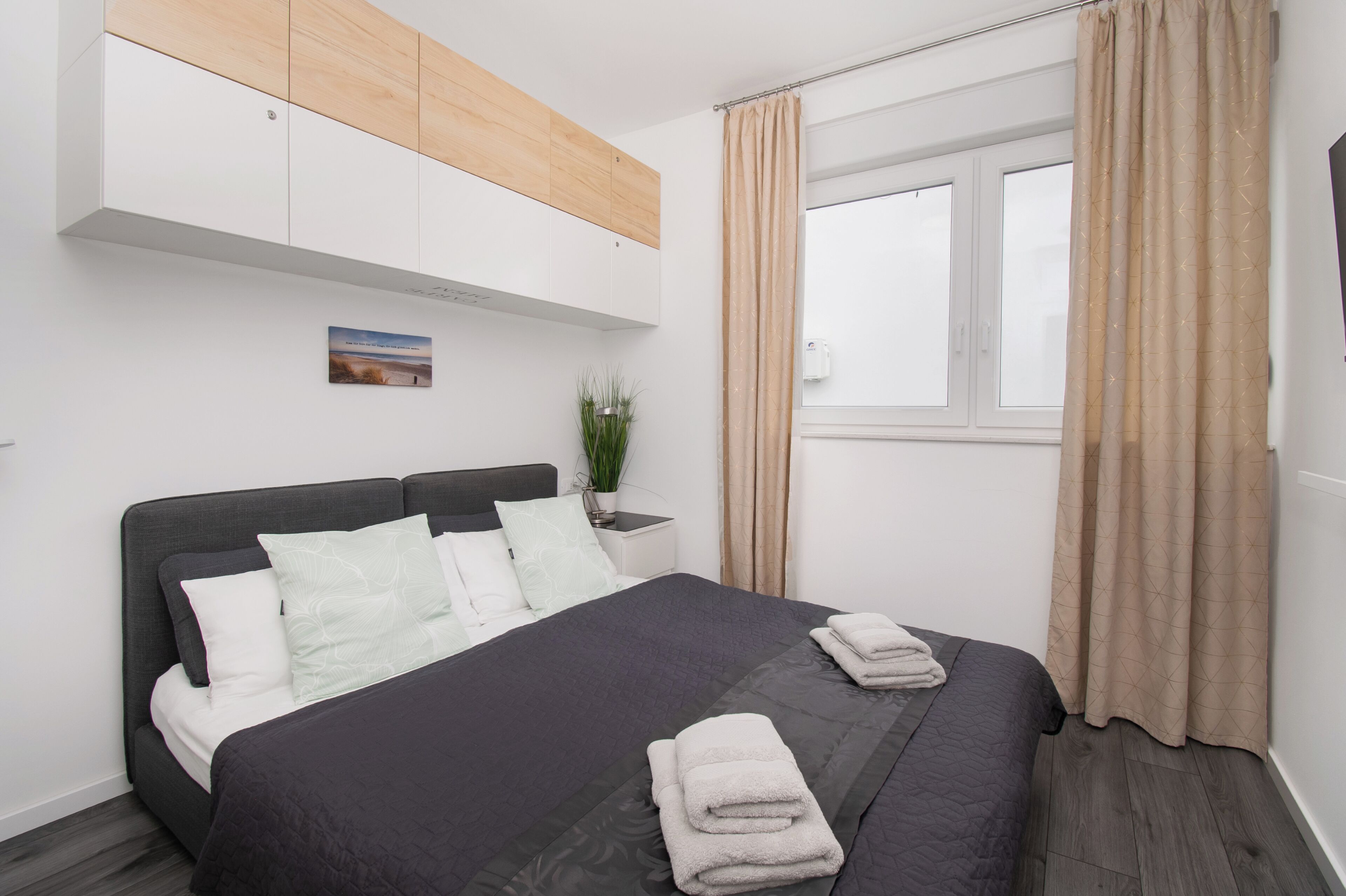 A modern bedroom with a bed, a nightstand, a potted plant, and a window with curtains.