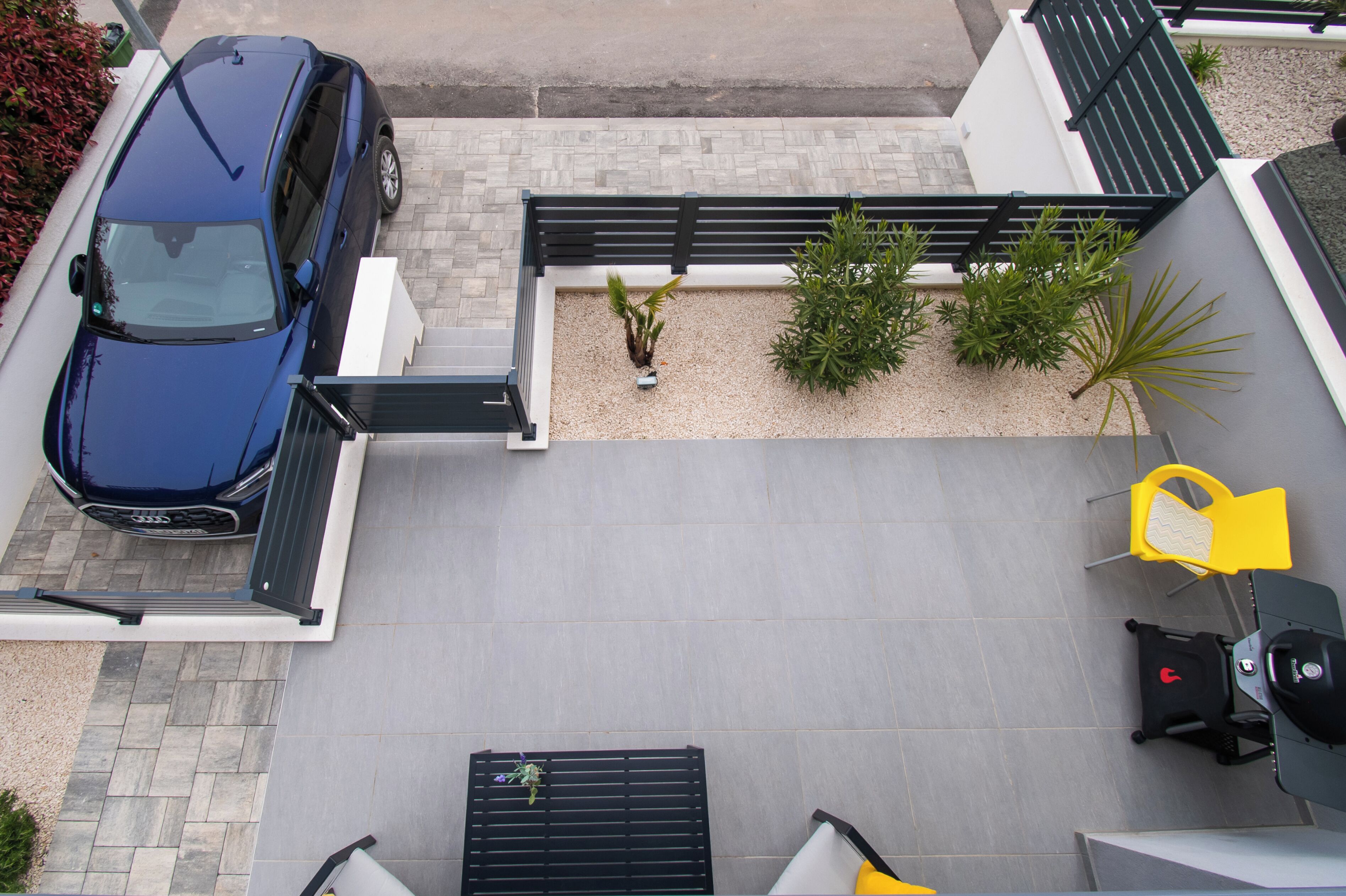 Un espacio exterior moderno con un coche azul estacionado, un pequeño jardín con plantas y un área de descanso con sillas amarillas.