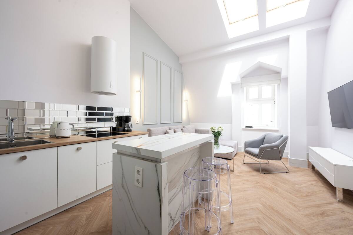 A modern kitchen with white cabinets, a marble island, and a dining area with a grey sofa and clear stools.