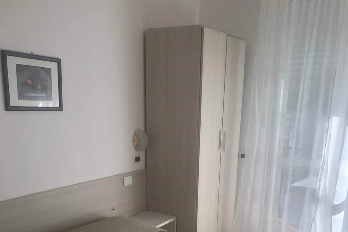 A hotel room with a bed, a wardrobe, a framed picture on the wall, and a window with sheer curtains.