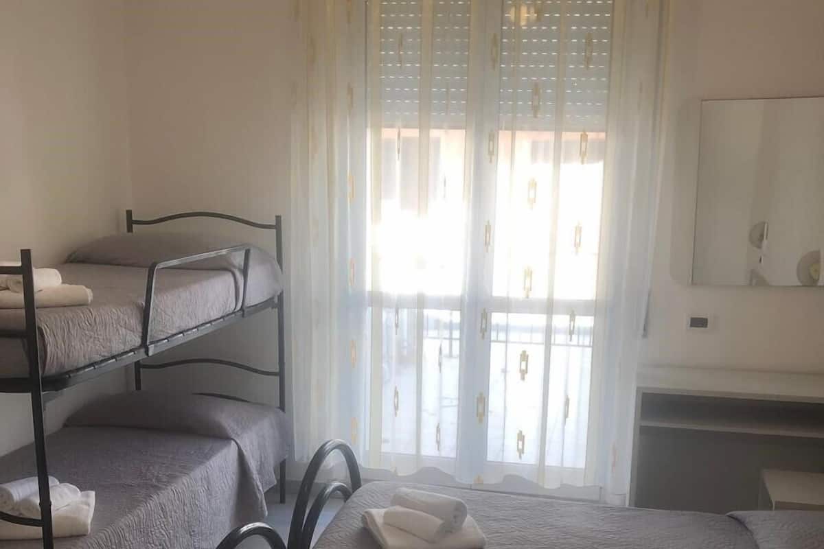 A dormitory room with bunk beds, a desk, and a window with sheer curtains.