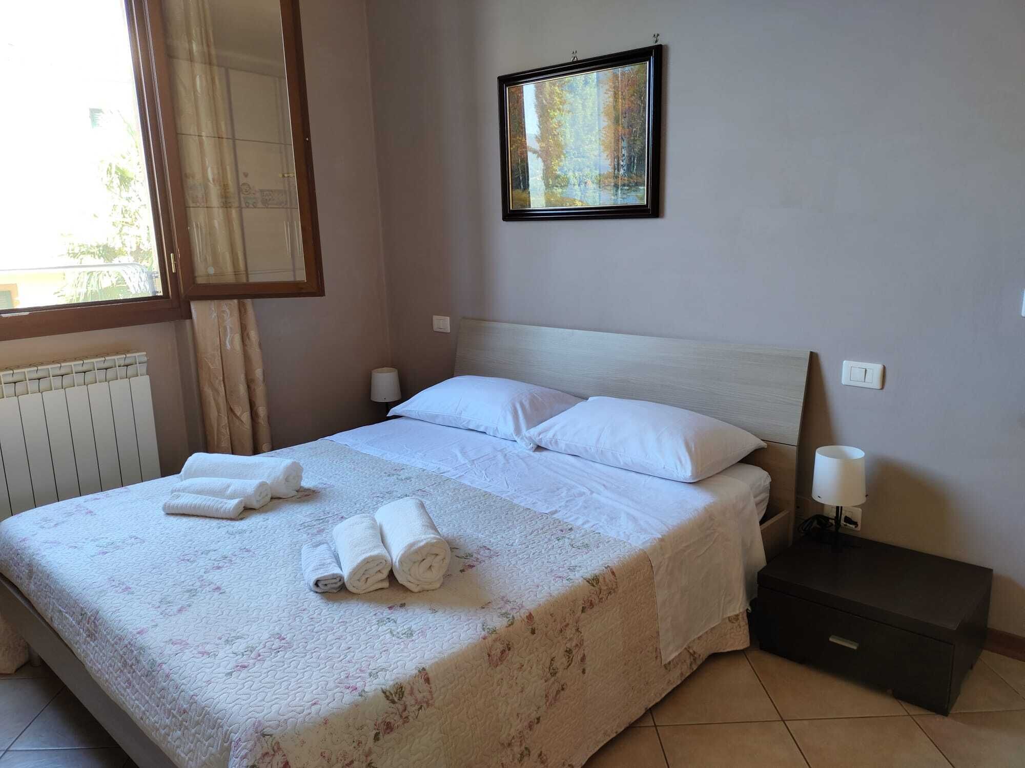 A neatly made bed with white linens and two rolled towels, a bedside table with a lamp, and a framed picture on the wall.