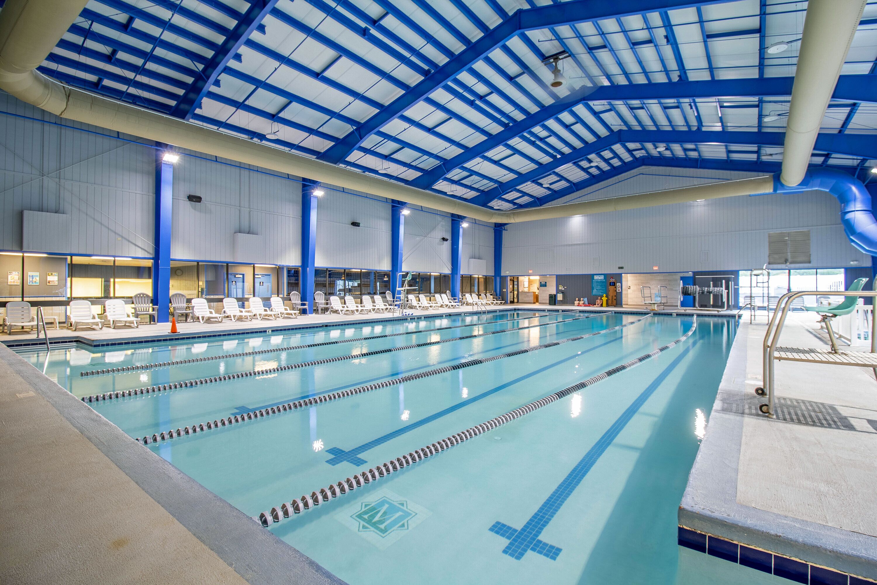 An indoor swimming pool with multiple lanes, lounge chairs, and a high ceiling with blue structural beams.