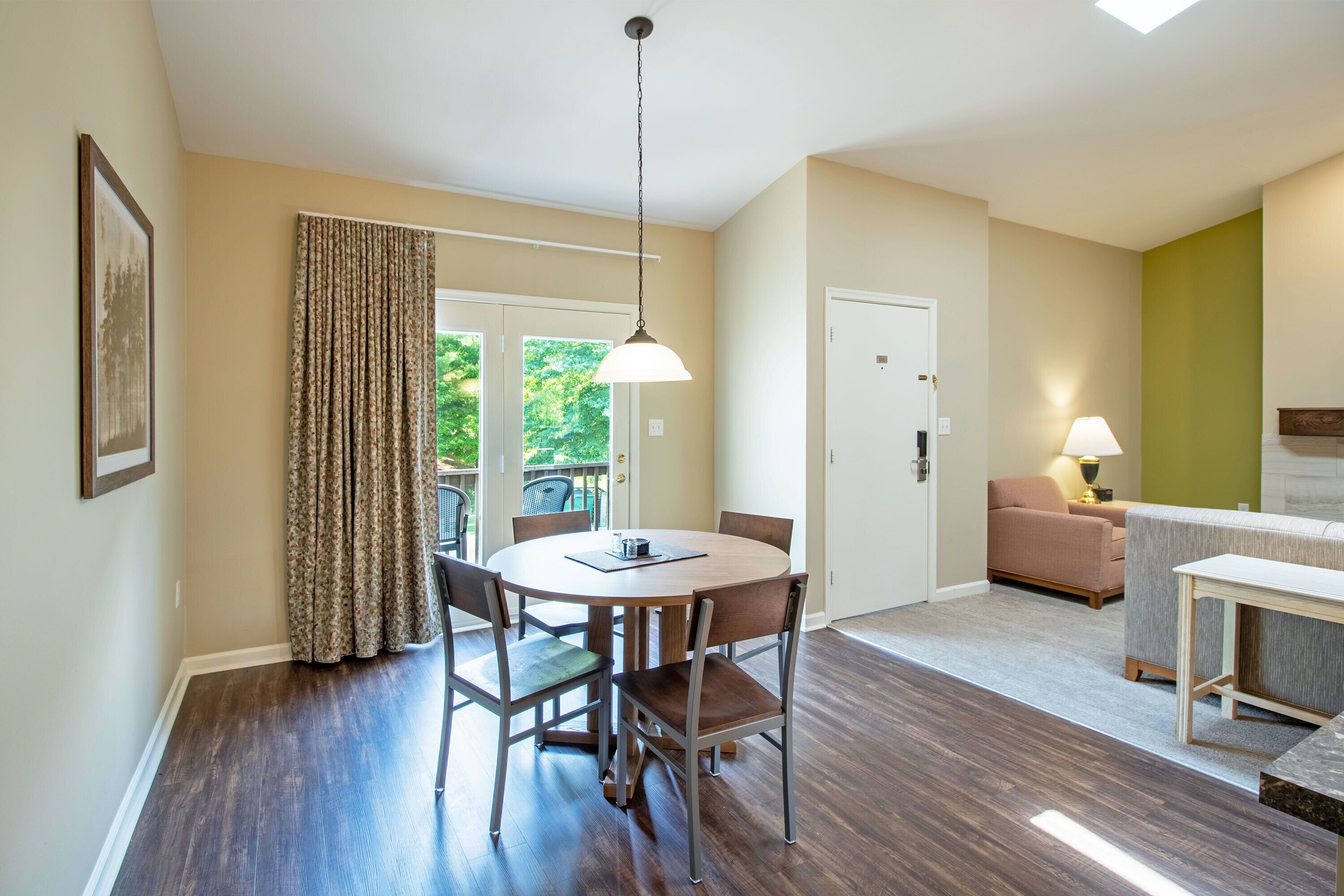 A dining area with a round table and chairs, a hanging light fixture, a door leading to a balcony, and a sofa in the adjacent living area.