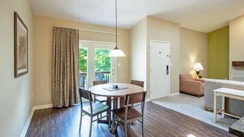 A dining area with a round table and chairs, a hanging light fixture, a door leading to a balcony, and a sofa in the adjacent living area.