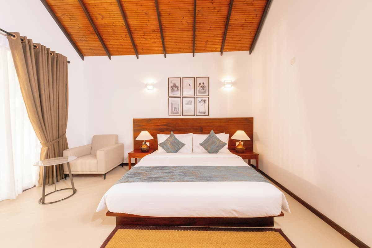Un dormitorio con cabecera de madera, cama con ropa de cama blanca, dos lámparas de noche, una mesita redonda pequeña, una silla y un aplique de pared.