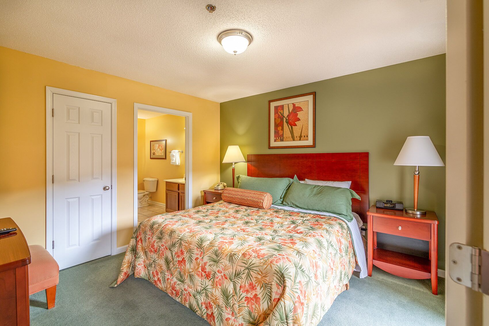 Habitación de hotel con cama, mesita de noche, lámpara, puerta y baño visible a través de una puerta abierta.