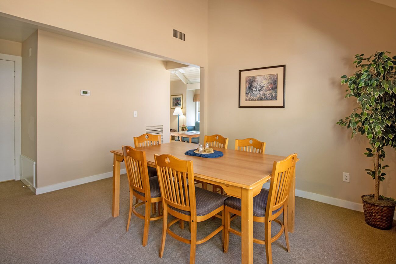 A dining area with a wooden table and chairs, a framed picture on the wall, and a potted plant.
