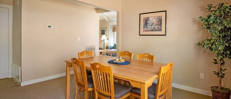 A dining area with a wooden table and chairs, a framed picture on the wall, and a potted plant.