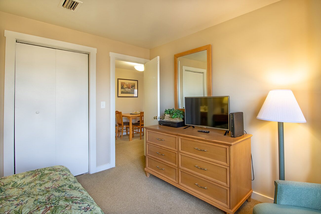 Un dormitorio con cama, cómoda, televisión, lámpara y un área de comedor al fondo.