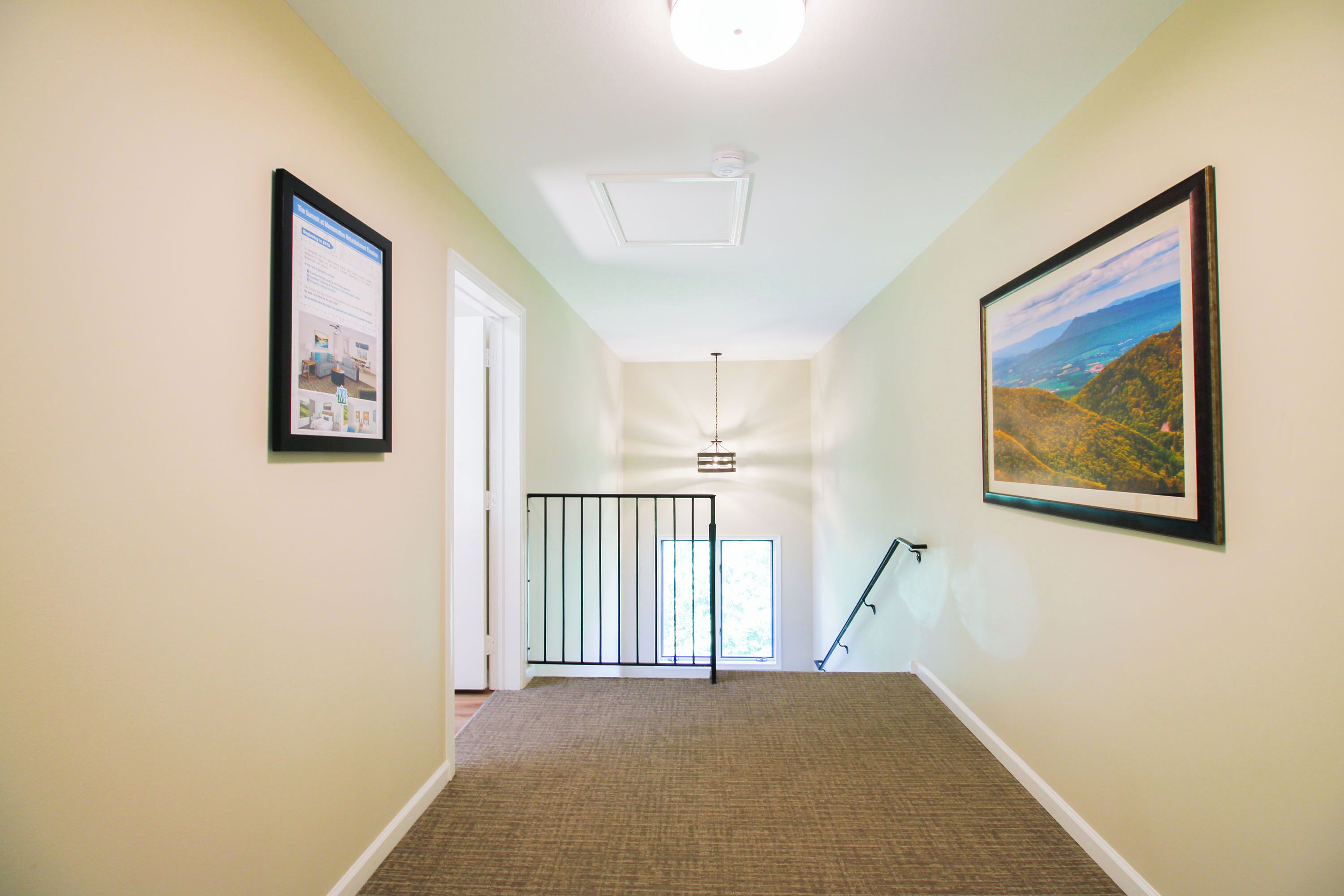 A hallway with beige walls, a carpeted floor, a ceiling fan, and framed pictures on the walls.