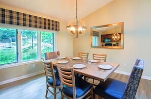 Une salle à manger avec une table dressée pour quatre personnes, des chaises, un lustre et une grande fenêtre donnant sur un paysage verdoyant.