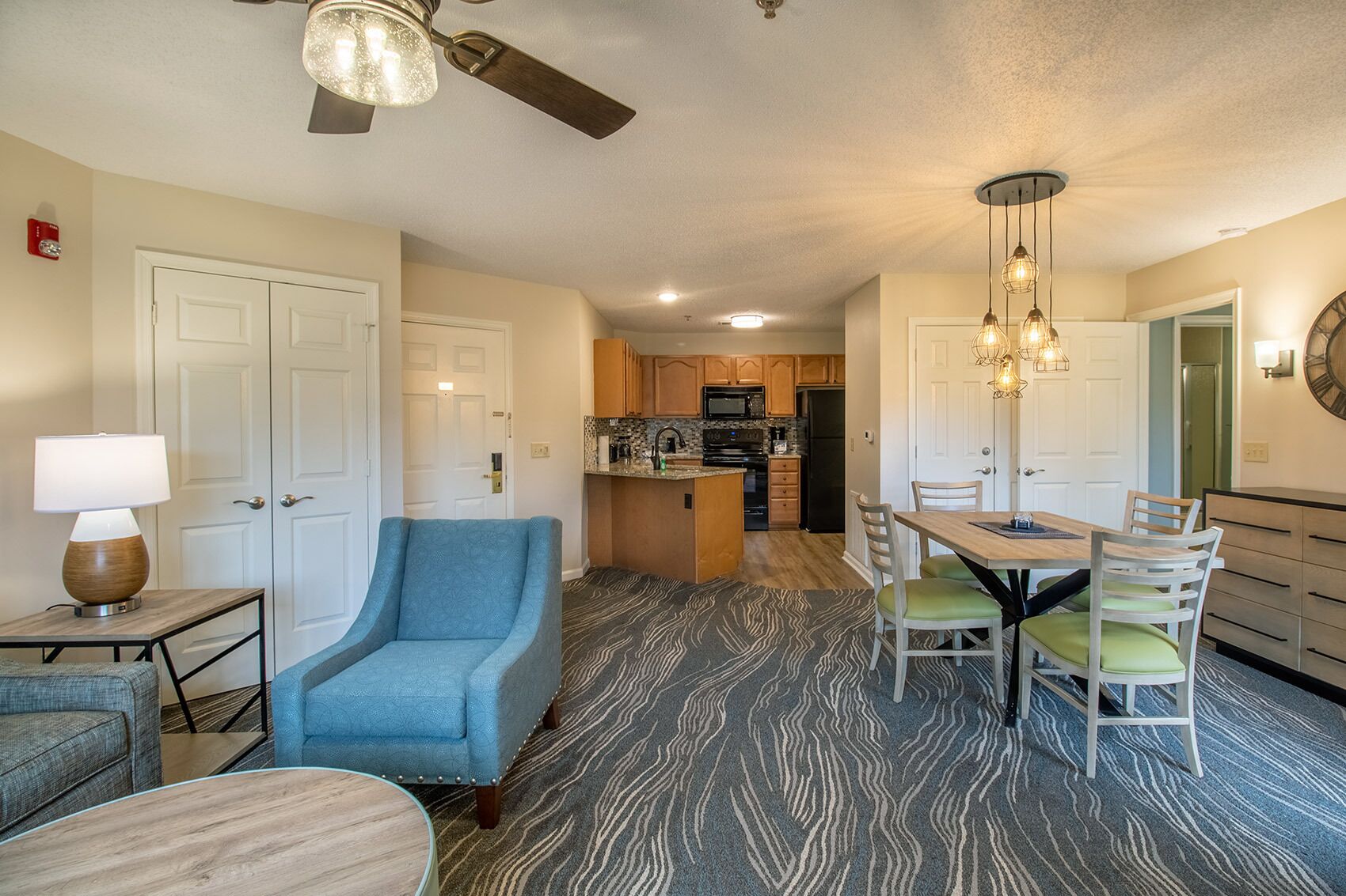 A compact living space with a kitchenette, a dining area with a table and chairs, a blue armchair, and a ceiling fan.