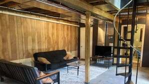 A modern living room with a black sofa, a glass coffee table, and a TV on a stand. There is a wooden wall and a concrete floor. - Riverview Retreat - house with sauna in the New River Gorge National Park! (Meadow Creek)