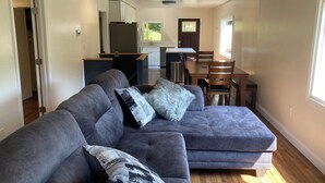 A modern living room with a sectional sofa, a dining area with a wooden table and chairs, and a kitchen with white cabinets and a refrigerator. - Riverview Retreat - house with sauna in the New River Gorge National Park! (Meadow Creek)