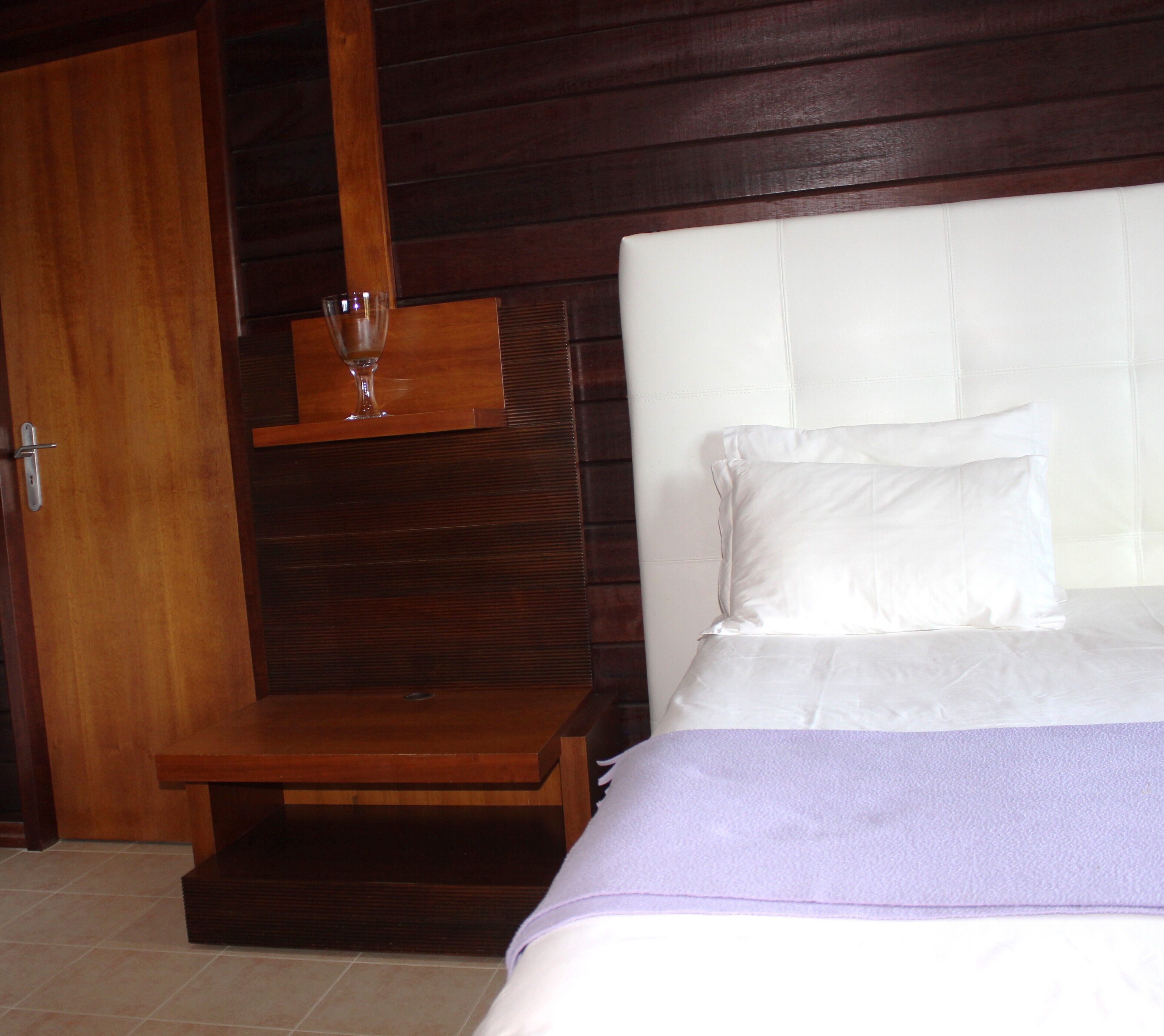 A bedroom with a wooden headboard, a bed with white bedding, and a small wooden nightstand with a glass on it.