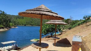 Zona de playa con dos sombrillas de paja, una mesa de madera, un bote pequeño y una tabla de surf.