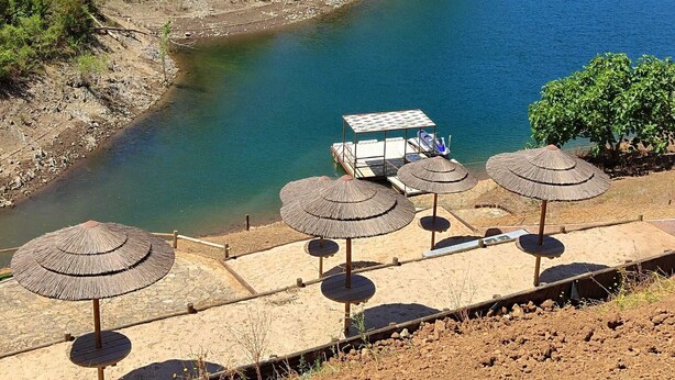 Vista al río con un muelle de madera, sombrillas de paja y un barco pequeño.