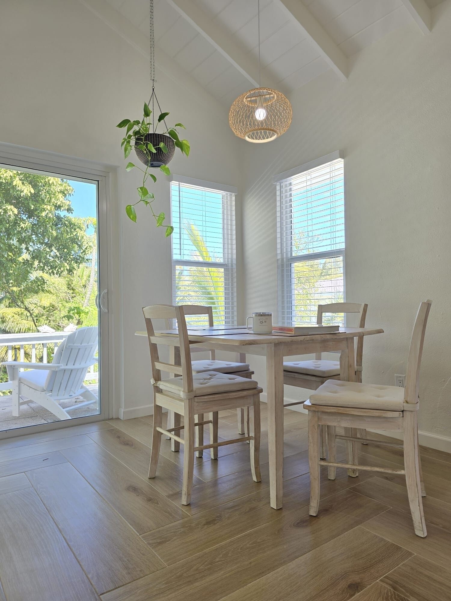 A dining area with a wooden table and chairs, a hanging plant, and large windows with blinds.