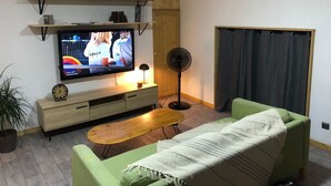 A living room with a TV, wooden shelves, a fan, a sofa, a coffee table, and a potted plant.