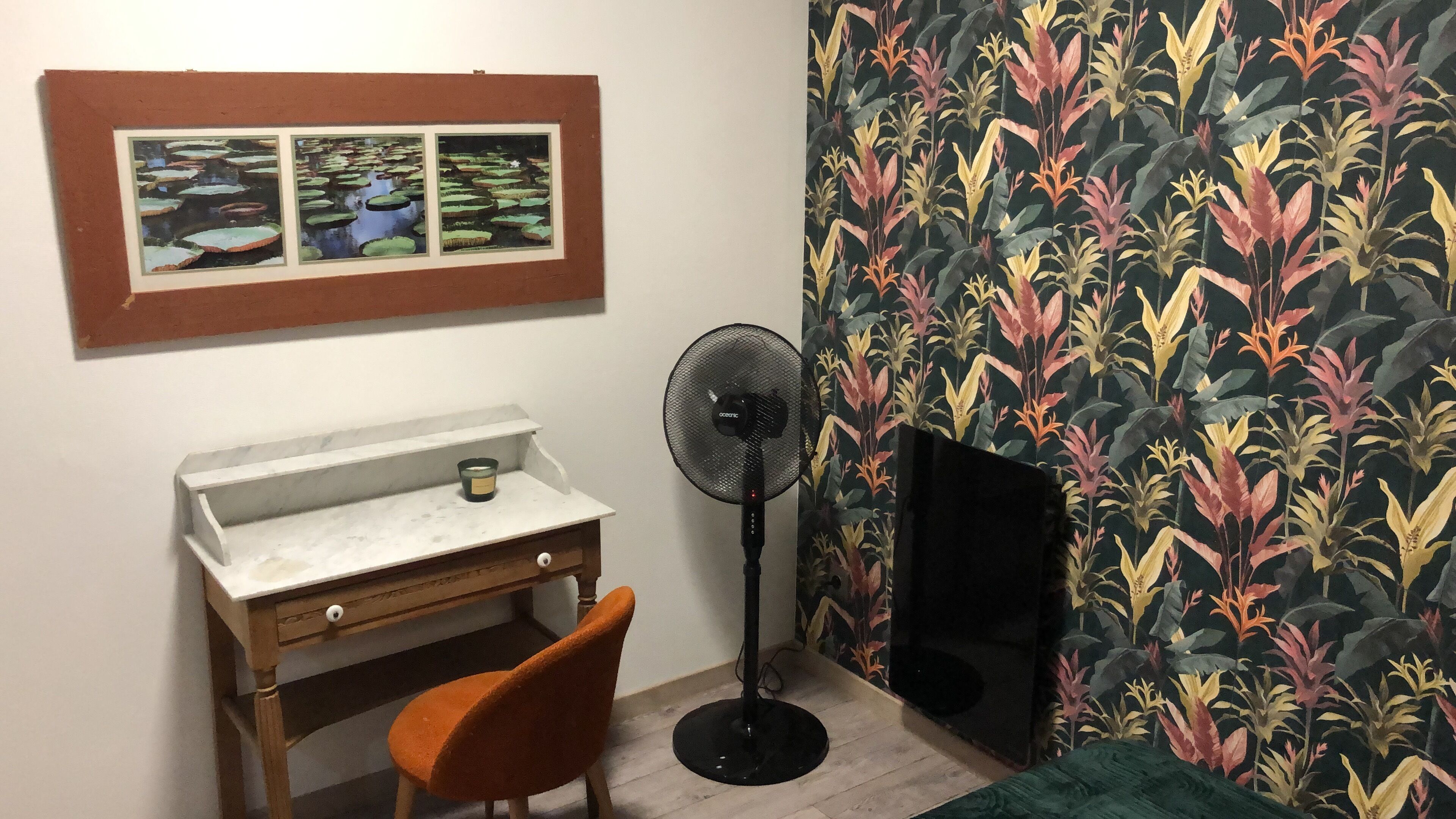 Quarto com escrivaninha, cadeira e ventoinha. Há um quadro na parede e um papel de parede estampado.
